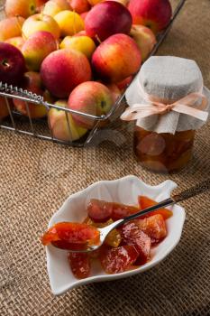Homemade slices of apple jam on white saucer, spoon, jar of marmalade and apples in metal wire basket on a burlap covered table, vertical. Selective focus. The toning