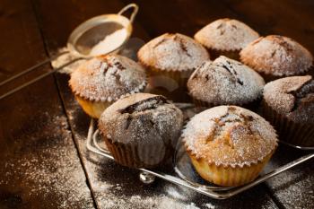 Vanilla and chocolate muffins on metal stand, caster sugar, sieve for a baking on a dark wooden background, selective focus