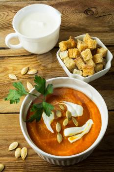 Pumpkin squash cream soup with cream and pumpkin seeds in the white bowl, cream in a creamer and croutons in a serving dish on the dark wooden background, vertical