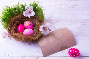 pink Easter eggs in a nest with white flowers in the green fresh grass on the white wooden background with natural unbleached paper for congratulation. Easter background. Easter symbol. Copy space