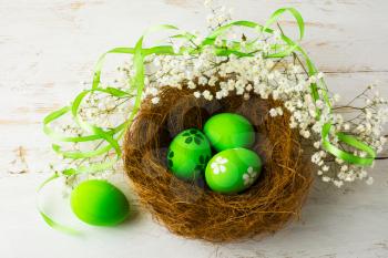Green Easter eggs in a nest with green satin ribbon and small white baby's breath flowers on a white wooden background, top view