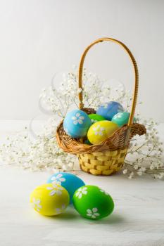 Decorated light blue, yellow, green Easter eggs in the basket with small white baby's breath flowers on a white wooden background, vertical