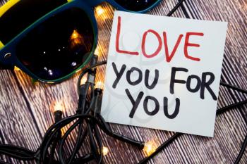 Love You For You handwritten on white note paper and love written in red color and further written in black color. Two Glasses with wooden background and lights. Valentine day concept. 