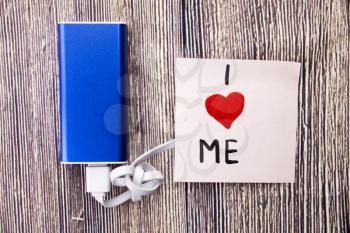 I love me heart handwritten on note paper in different colors and heart shape in red color. Energy storage device of blue color with cable on wooden background. selfish or self love Concept.