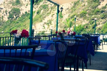 Restaurant tables with black chairs red and white flowers and blue tablecloth in natural environment. Peaceful place to have food. Having meal on table with great view