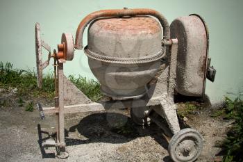 Dirty mini cement mixer in the the ground with little rocks and stones , tall green grasses behind it. Newly used equipment to build, fix structure put at the side of the house after done. 