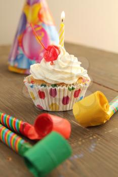 Vanilla cupcake with white frosting, cherry on top and a candle with party hat and blowers for birthday party 