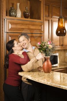 Caucasian woman kissing Caucasian man on cheek as he arranges flowers in vase in kitchen.
