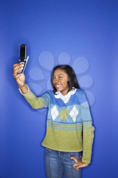 Portrait of African-American teen girl taking photo with camera phone standing in front of blue background.
