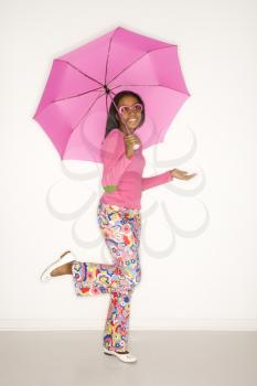Portrait of African-American teen girl holding a pink umbrella with leg bent behind her and holding hand out checking for rain in front of white background.