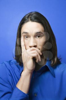 Portrait of Asian-American teen boy with hand covering chin and finger on side of face against blue background.
