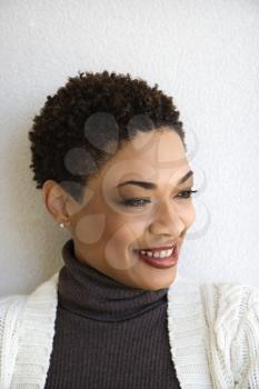 Close up head and shoulder of African-American woman standing against white wall smiling with head turned to side.