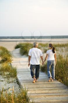 Back view of mid-adult Caucasian couple walking down walkway to beach.