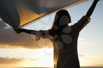 Royalty Free Photo of a Woman Holding Up Fabric in the Breeze Silhouetted by Sunset Beside Ocean