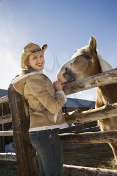Young Caucasian woman standing at fence with horse looking at viewer.