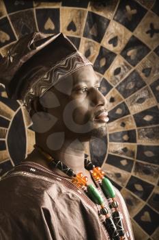 Profile portrait of an African American man wearing traditional African clothing, in front of a patterned wall. Vertical format.