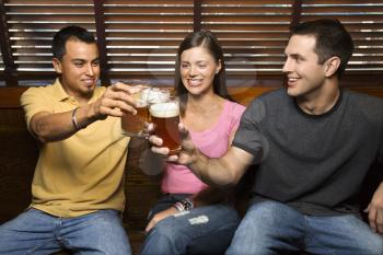 Three smiling young adult friends sitting on a bench toasting with their beers. Horizontal shot.