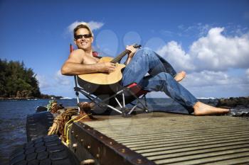 Shirtless young man wearing sunglasses, sitting on a pier and playing a guitar and smiling. Horizontal shot.