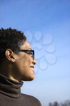 Attractive young African American woman in glasses smiles and looks out to the side during a sunny blue day. Vertical shot.