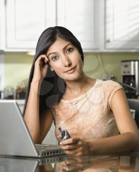 Young woman looks towards the camera while sitting in front of a laptop and holding a camera. She is leaning her head on her hand. Vertical shot.