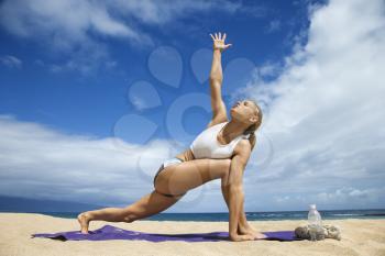 Attractive young woman extends an arm while and doing yoga on beach with the ocean in the background. Horizontal shot.