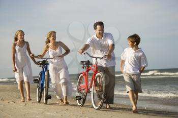 Family walks down the beach as mother and father push their bicycles. Horizontal shot.
