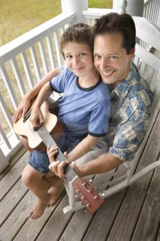 Boy playing guitar while sitting on his father's lap. Vertical shot.