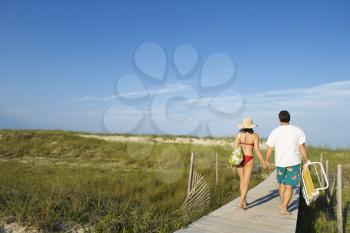 Man and woman wearing swimwear hold hands and walk down a boardwalk. Horizontal shot.