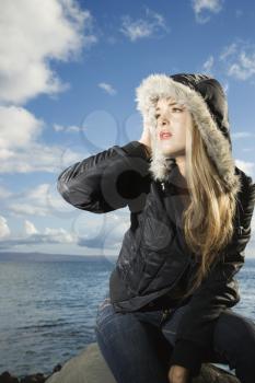 Attractive young woman in a parka with her with hand on her head sitting on a rock at the beach. Vertical shot.