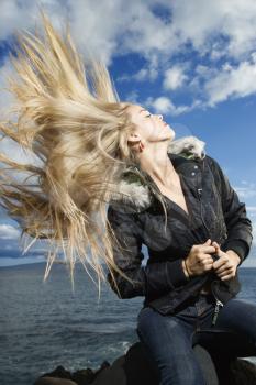 Attractive young woman sitting on a rock at the beach and flipping her long blond hair with the ocean in the background. Vertical shot.