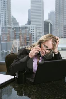 Caucasian businesswoman on the phone at her desk, holding her head and looking at a laptop computer with a frustrated expression. City buildings can be seen in the background through the window. Verti