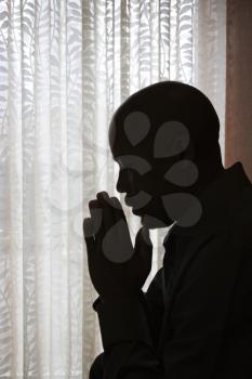 Profile of African-American man silhouetted by a window with his head bowed and his hands in prayer. Vertical shot.
