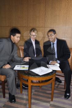 Three ethnically diverse business people sit at a small table and look at a laptop together. Vertical format.