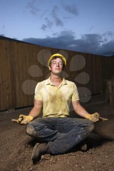 Caucasian male construction worker sits in a yoga meditation pose while wearing a yellow hardhat. Vertical shot.