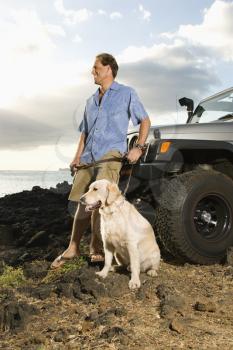 A smiling man leans against his SUV as he holds his dog's leash and both look off into the distance at a beach. Vertical format.