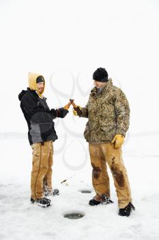 Two young men clink beers bottles while ice fishing. Vertical shot.