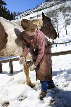 Man wearing a cowboy hat cleans out a horse hoof in the snow. Vertical shot.