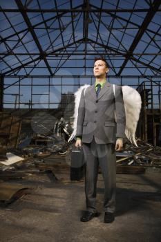 Serious young businessman wearing angel wings holds a briefcase inside an abandoned building. Vertical shot.