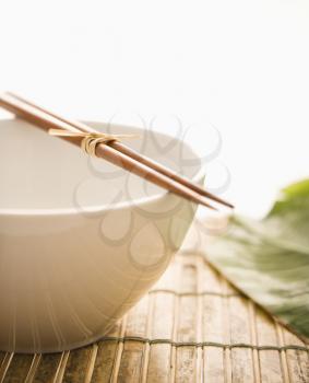 Chopsticks lying across an empty bowl on a bamboo mat, with a green leaf in the background. Vertical format. Isolated on white.