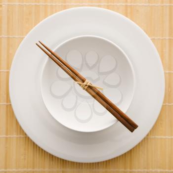 Overhead view of chopsticks lying across an empty bowl that is sitting on a plate on a bamboo mat. The dishes are white. Square shot.