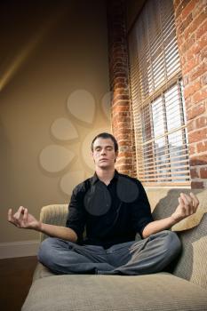 Young Caucasian man sitting on living room sofa in lotus pose.