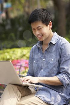 Young Chinese Man Using Laptop Whilst  Relaxing On Park Bench