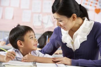 Teacher Helping Student Working At Desk In Chinese School Classroom