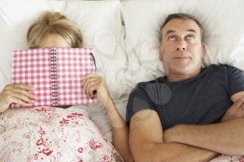 Bored Looking Senior Man Lying In Bed Next To Senior Woman Reading Book
