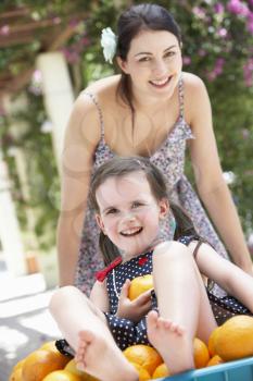 Mother Pushing Daughter In Wheelbarrow Filled With Oranges