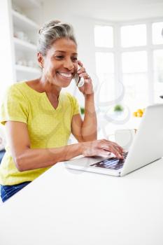 Woman Using Laptop And Talking On Phone In Kitchen At Home