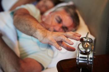 Couple In Bed With Man Reaching To Switch Off Alarm Clock