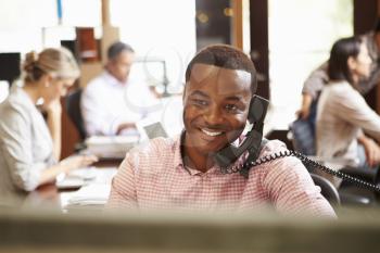 Businessman On Phone At Desk With Meeting In Background