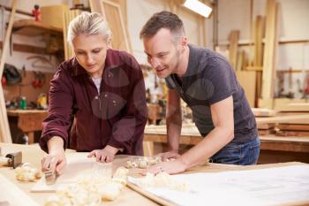 Carpenter With Female Apprentice Planing Wood In Workshop