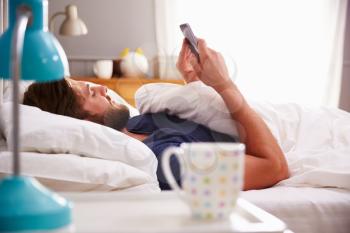 Man Lying In Bed Checking Mobile Phone